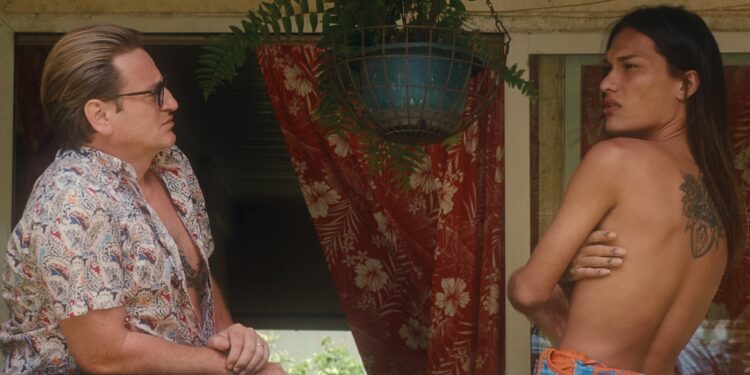 A photo of a scene from the film Pacifiction. Actors Benoît Magimel and Pahoa Mahagafanau are looking at each other on some decking stood in front of a red curtain