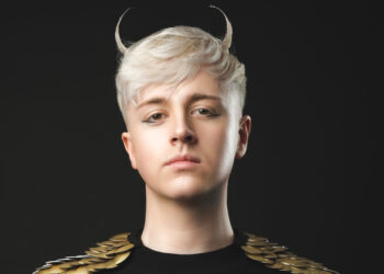 A photo of comedian Daniel Fox wearing a black top with gold sleeves. Their short blonde hair has a sweeping fringe and is fashioned in to Devil horns at the top