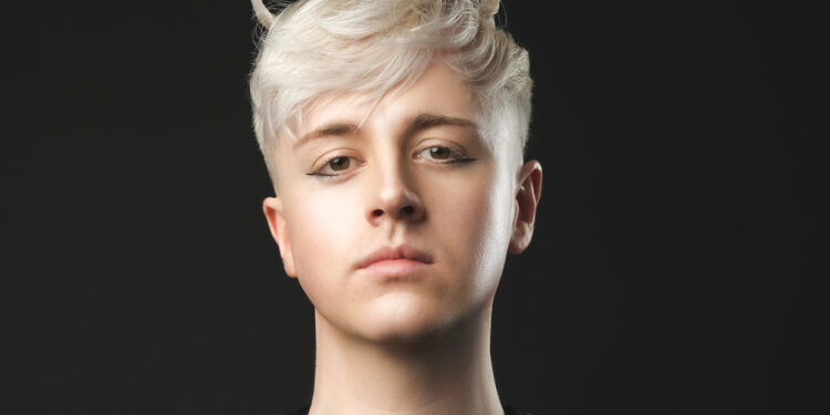 A photo of comedian Daniel Fox wearing a black top with gold sleeves. Their short blonde hair has a sweeping fringe and is fashioned in to Devil horns at the top