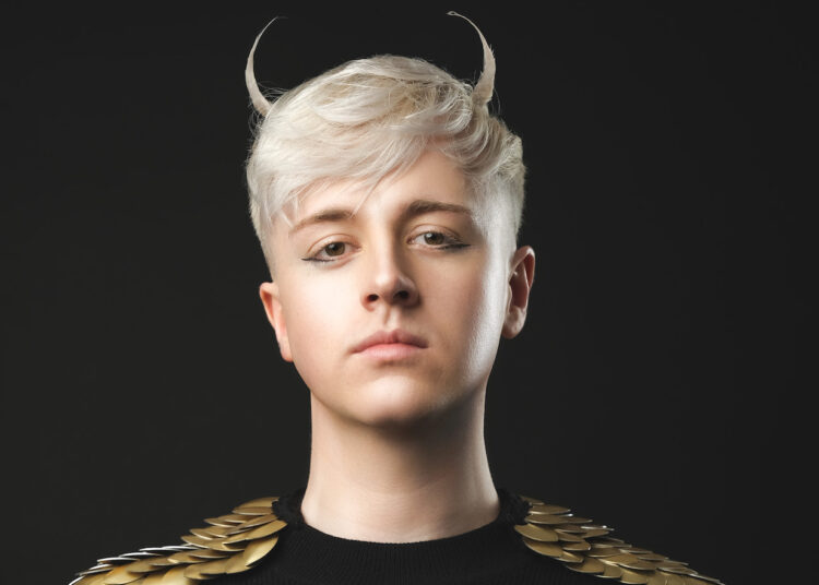 A photo of comedian Daniel Fox wearing a black top with gold sleeves. Their short blonde hair has a sweeping fringe and is fashioned in to Devil horns at the top