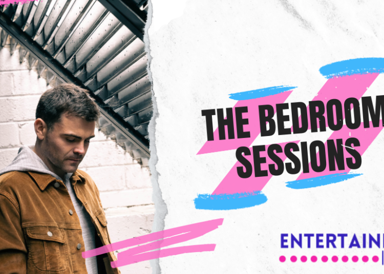 A photo of a man with dark hair with a brown jacket and a green hooded top underneath. he is looking down at the floor and is stood outside in front of a white brick wall