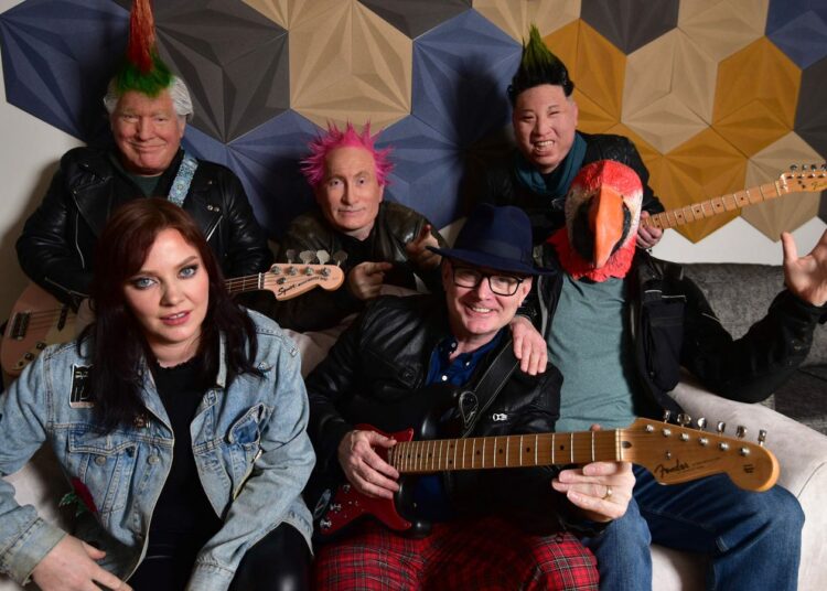 The band Beldon Haigh are posing for a promo shot on a sofa they are a 6 piece band and 3 of them are wearing masks of people and one is wearing a mask of a parrot