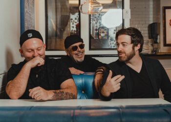 The band Big Yard sit at a table, there are three members and they are talking and laughing