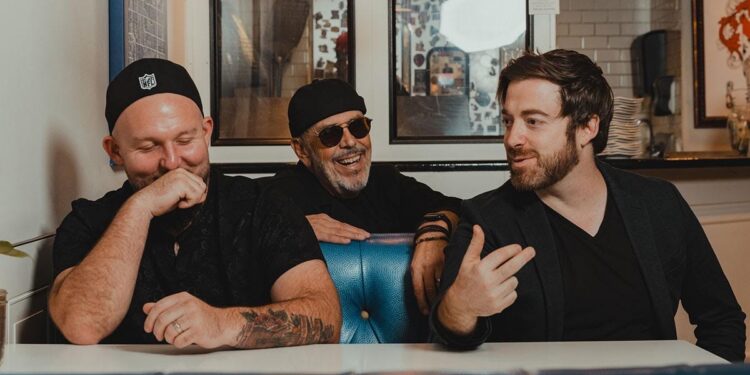 The band Big Yard sit at a table, there are three members and they are talking and laughing