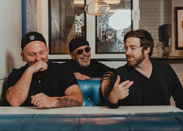 The band Big Yard sit at a table, there are three members and they are talking and laughing