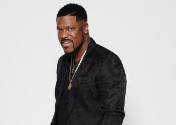 Actor and comedian Chris Tucker is stood side on and looking into the camera, he is wearing a black shirt with a gold necklace