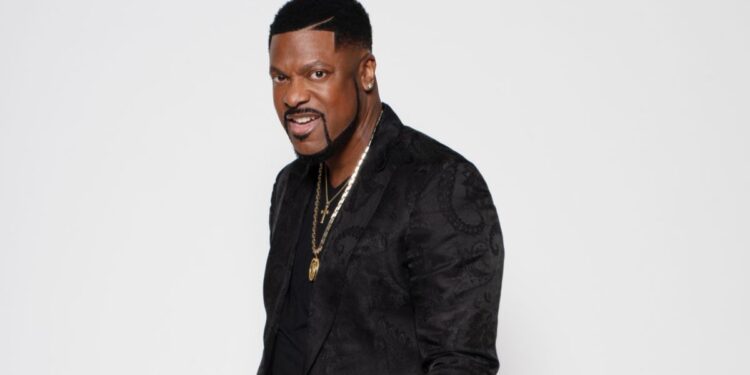 Actor and comedian Chris Tucker is stood side on and looking into the camera, he is wearing a black shirt with a gold necklace