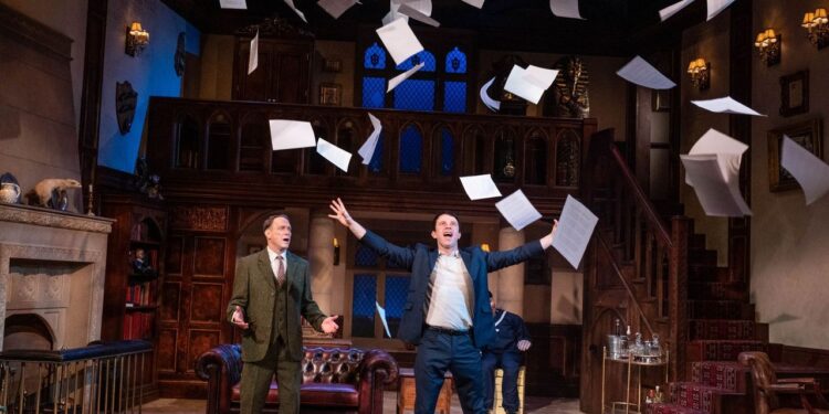 A photo of 2 male actors on stage, one is wearing a blue suit and throwing papers in to the air and the other is wearing a tweed suit and is looking shocked