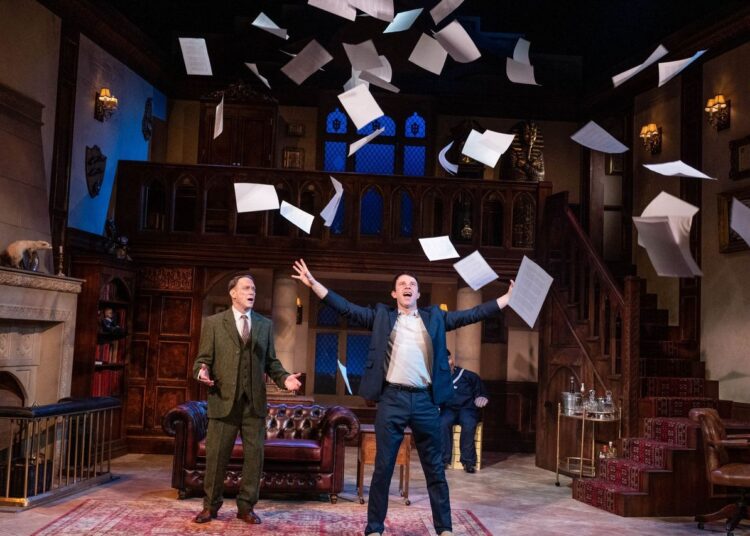 A photo of 2 male actors on stage, one is wearing a blue suit and throwing papers in to the air and the other is wearing a tweed suit and is looking shocked