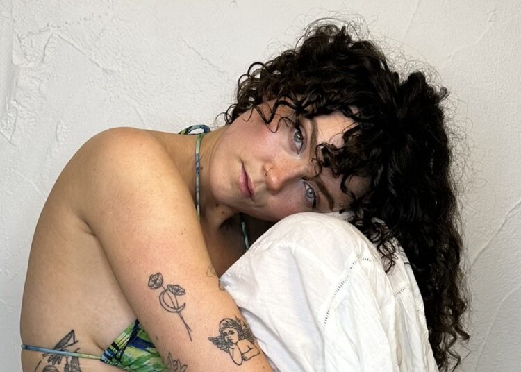 aakira fae sits with her head resting on her knees, she is wearing white trousers and a green bikini top