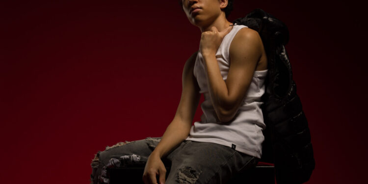 A young man poses seated in a dark red studio, wearing a white tank top and ripped jeans, with a small keyboard controller by his feet.