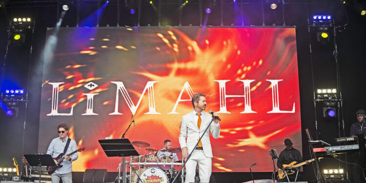 A live band performs on stage with a large screen behind them displaying the name “Limahl,” while the singer stands at the front holding a microphone.
