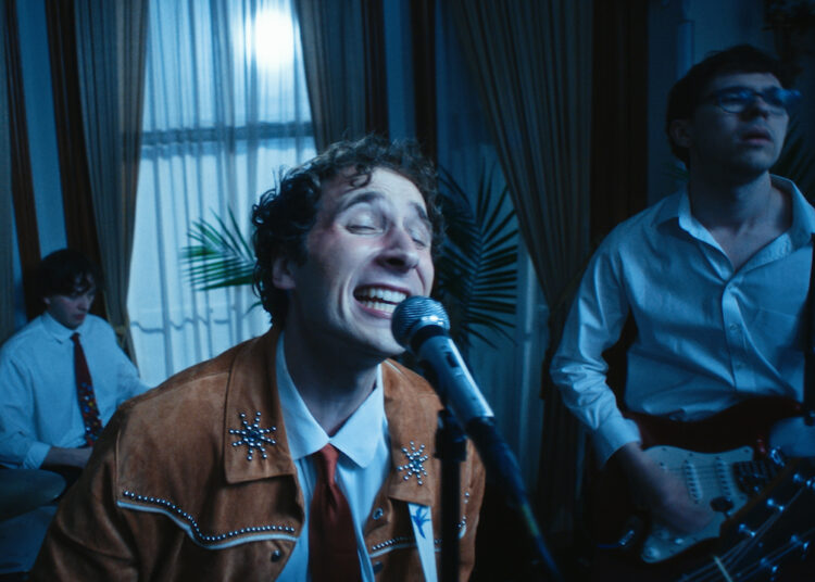 Peter DiMaggio performs indoors, with a band, singing into a microphone and musicians playing guitar and drums behind him