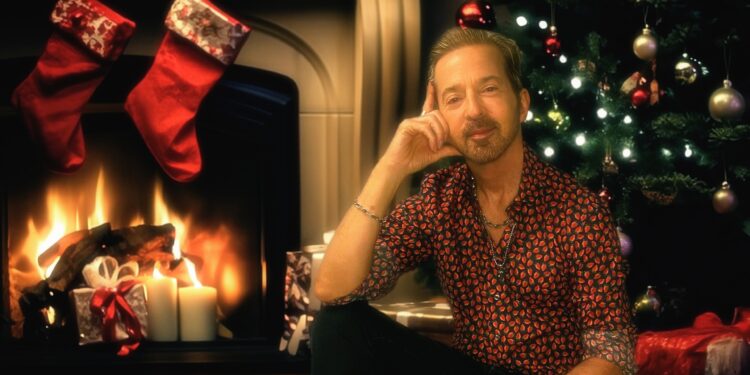Limahl sits by a fireplace and Christmas tree, smiling warmly in a festive room with stockings, candles, and wrapped gifts