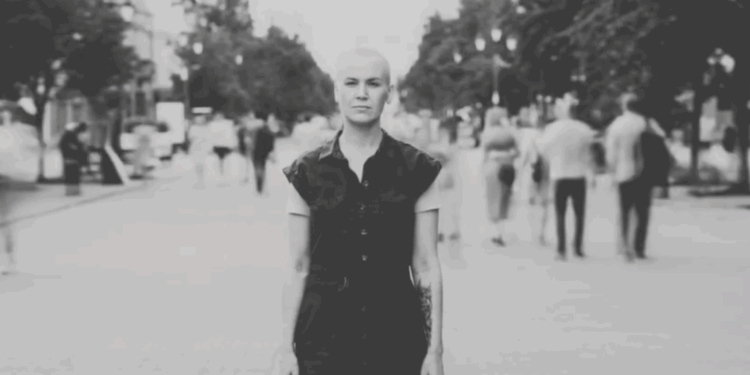 Black-and-white image of a woman standing still on a busy pedestrian street, with blurred people moving around her and the text “Time Stands Still – Gitika Partington” overlaid.