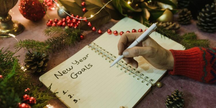 A hand writes “New Year Goals” in a notebook surrounded by festive decorations, pine branches and fairy lights.