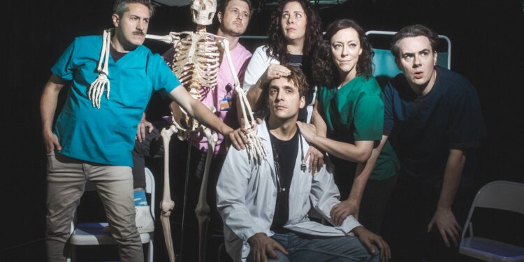 Group of performers in medical scrubs and lab coats posing dramatically around a seated man, with a skeleton prop under stage lighting.
