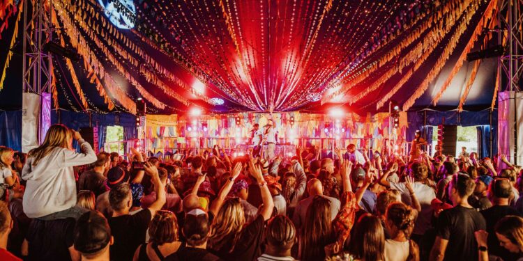Crowded festival tent with a live performance on stage, colourful bunting overhead and audience members raising their hands at Big Feastival