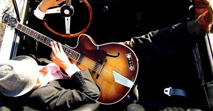 Overhead view of a musician sitting in the driver’s seat of a vintage convertible, holding a sunburst hollow-body guitar across his lap, with one hand on the steering wheel and sunlight illuminating the interior.