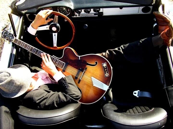 Overhead view of a musician sitting in the driver’s seat of a vintage convertible, holding a sunburst hollow-body guitar across his lap, with one hand on the steering wheel and sunlight illuminating the interior.